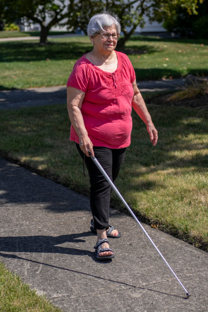 A woman in a red shirt and black pants walks down a sidewalk with her white cane.