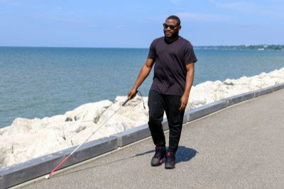 A man uses a white cane to walk down a paved path along lake Erie.