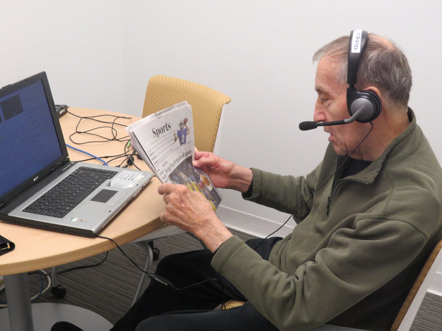 A radio reader reads aloud from the sports section of a printed newspaper. They're wearing a headset with a microphone that plugs into a laptop in the table in front of him.