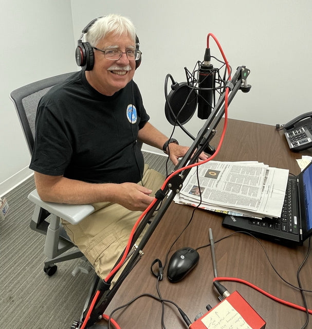 A radio reading volunteer smiles at the camera as a print newspaper sits on the table in front of him. A microphone is also set up in front of him. 