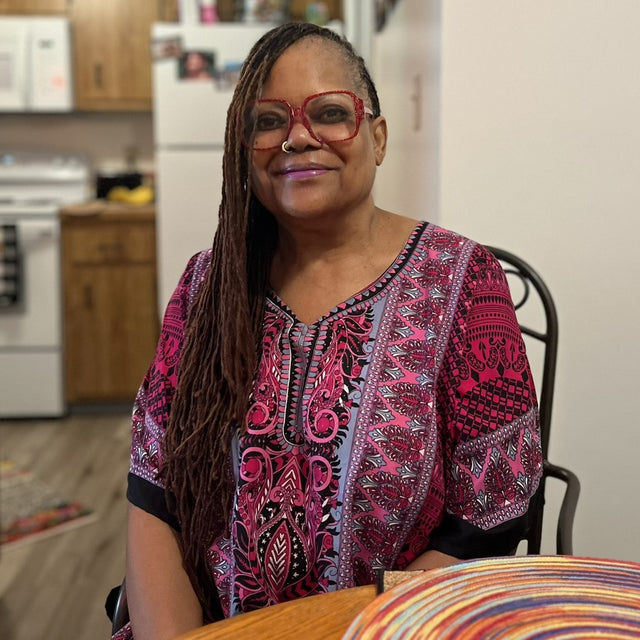 Rosie smiles in her home. She's wearing glasses and lipstick that match her purple and pink dress.